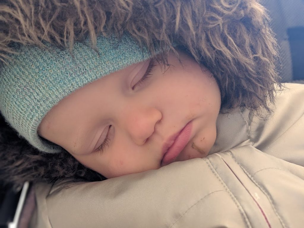 a toddler asleep in a stroller with a blue hat and a fur lined hood