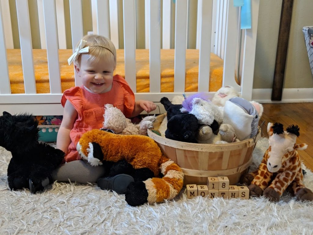 a girl toddler surrounded my all her stuffed animals in her room.