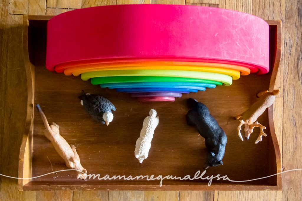 a Grimms rainbow stacker toy with forest animal figures in front of it on a wooden tray