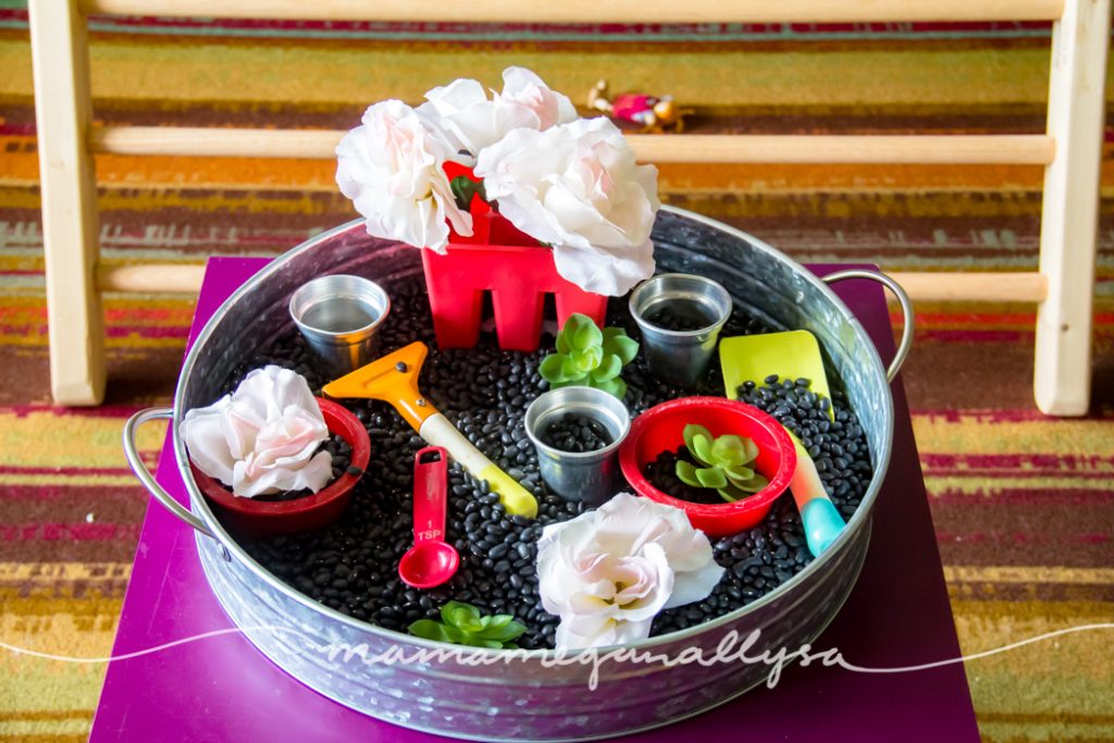 a black bean garden sensory bin containing red bowls, silver pots, fake flowers, and gardening tools