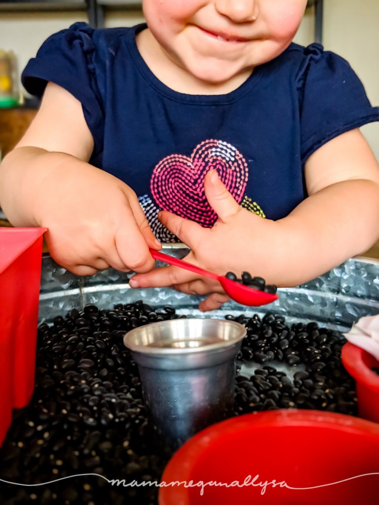 a toddler smiling holding a measuring spoon filled with black beans