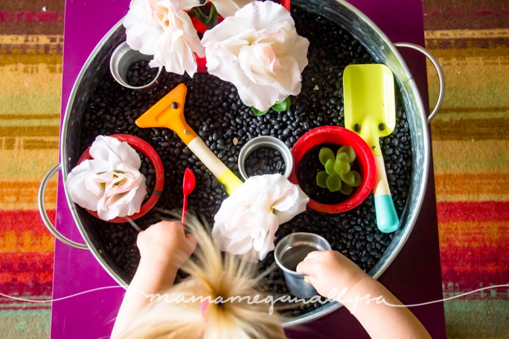 a top down view of toddler playing in a black bean garden sensory bin containing red bowls, silver pots, fake flowers, and gardening tools