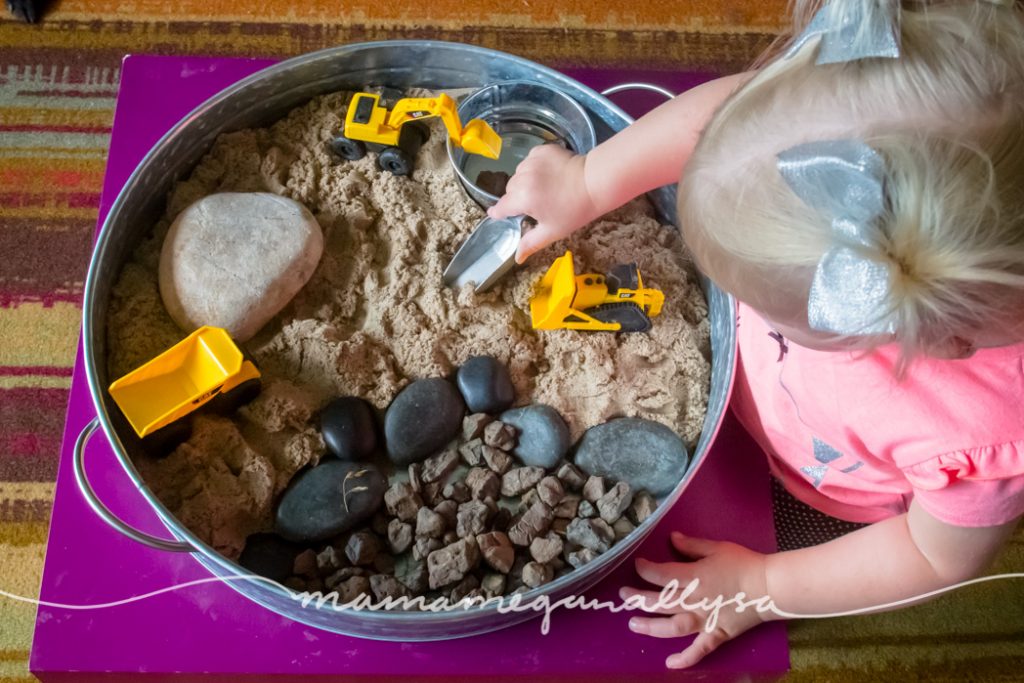 a toddler using a scoop in a tray of kinetic sand and stones