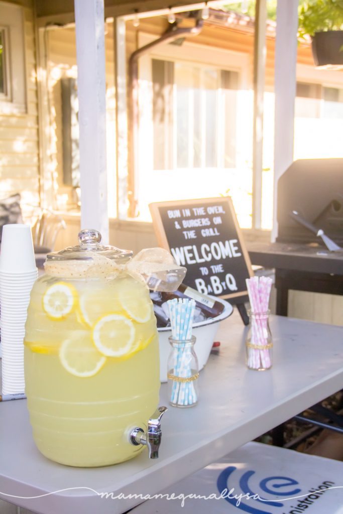 A dispenser of lemonade and some blue and pink paper straws in front of a letter board and the BBQ