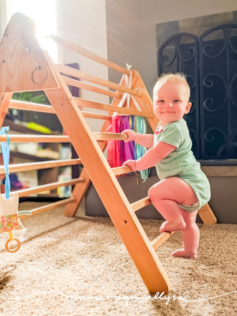 a baby with one foot up on a Pikler triangle (a wooden climbing toy shaped like a ladder folded in half)