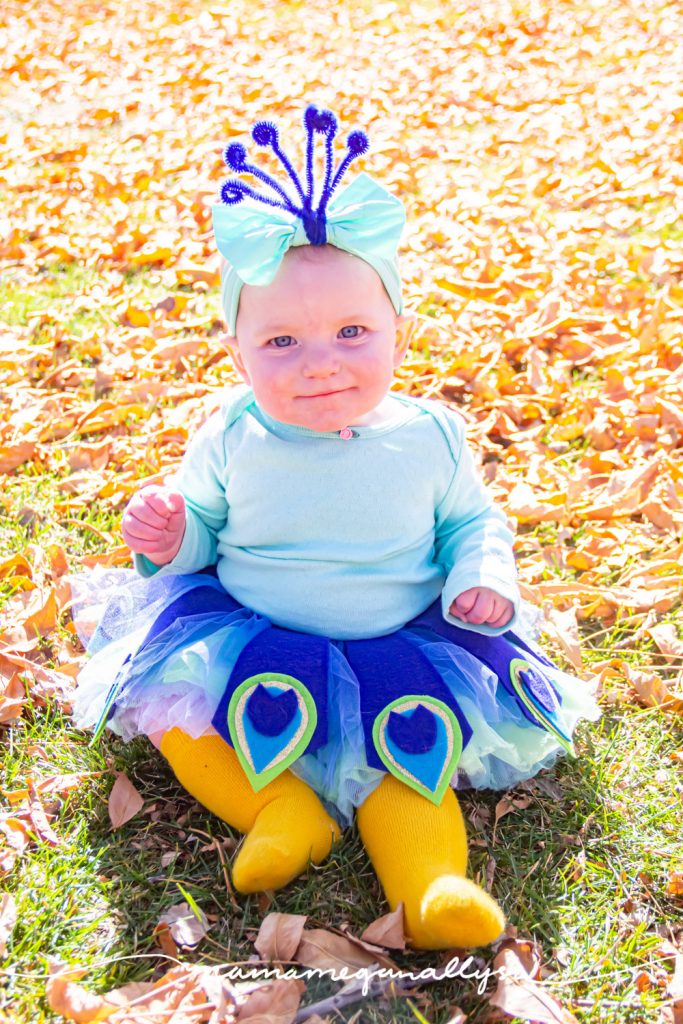 a baby dressed up like a homemade peacock costume for Halloween, sitting in the fall sunshine surrounded by leaves.