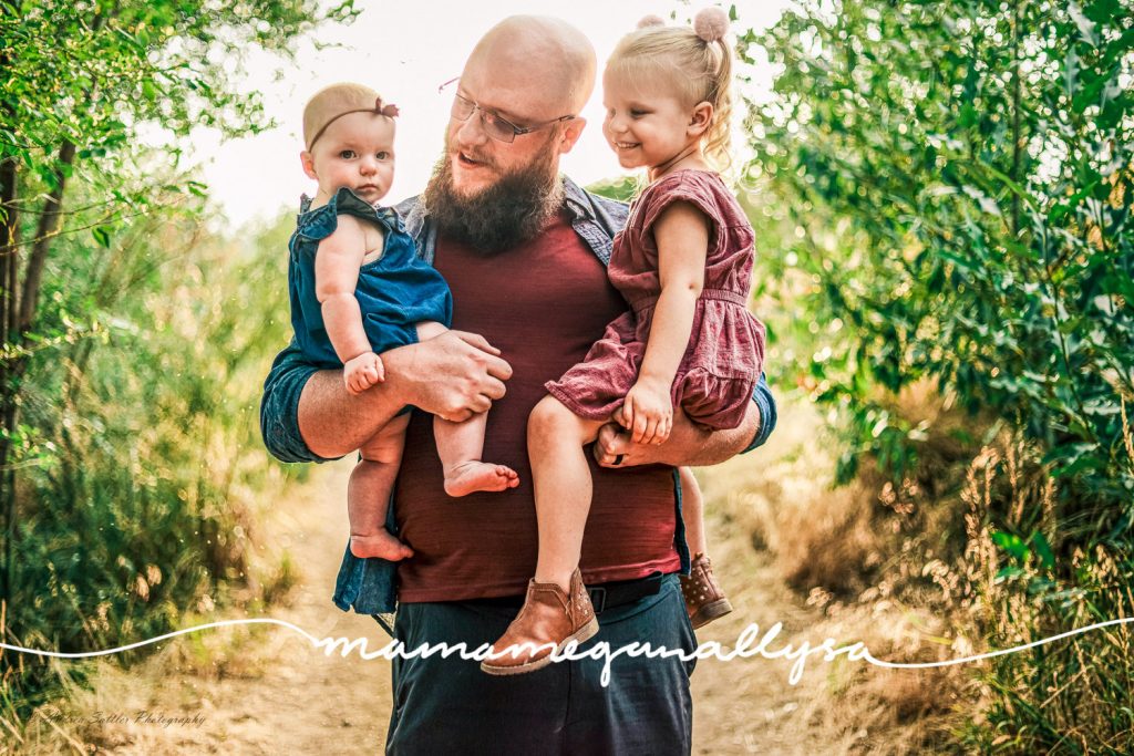a daddy holding both his girls in his arms. He and the toddler are looking at the baby and the baby is looking at the camera