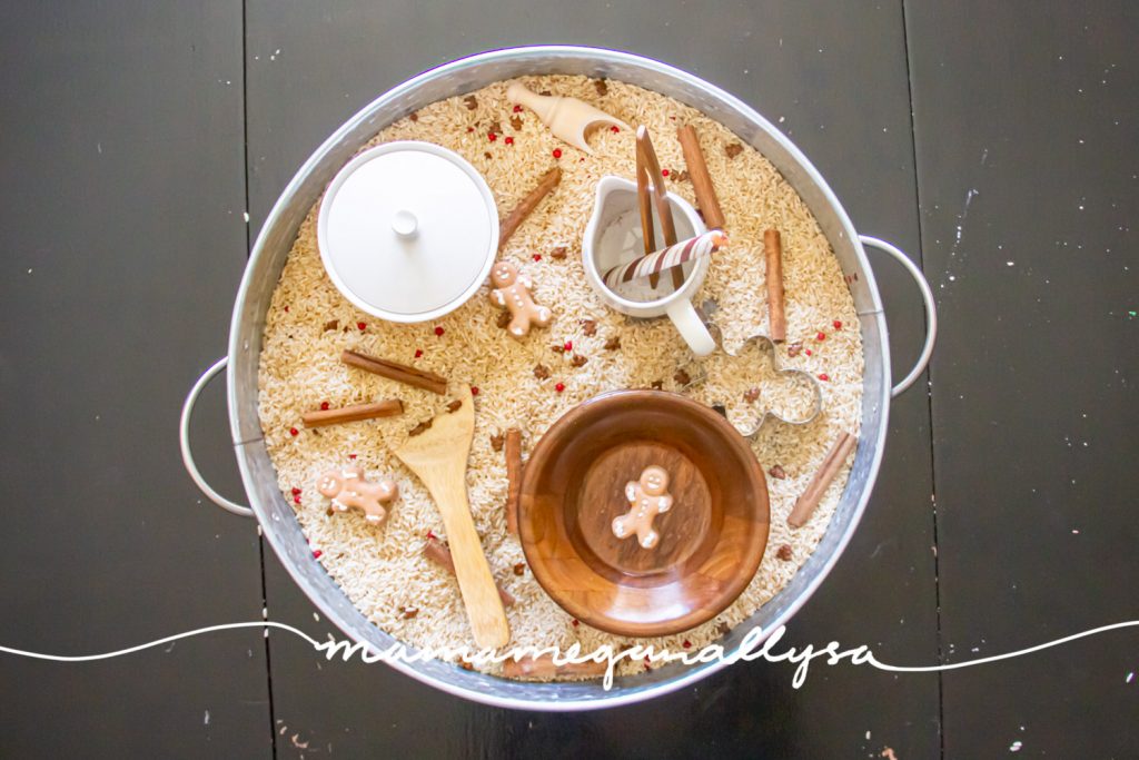 an overview of the setup gingerbread sensory bin. with the cinnamon sticks, sprinkles, and gingerbread men all mixed into the rice and the tolls scattered around.