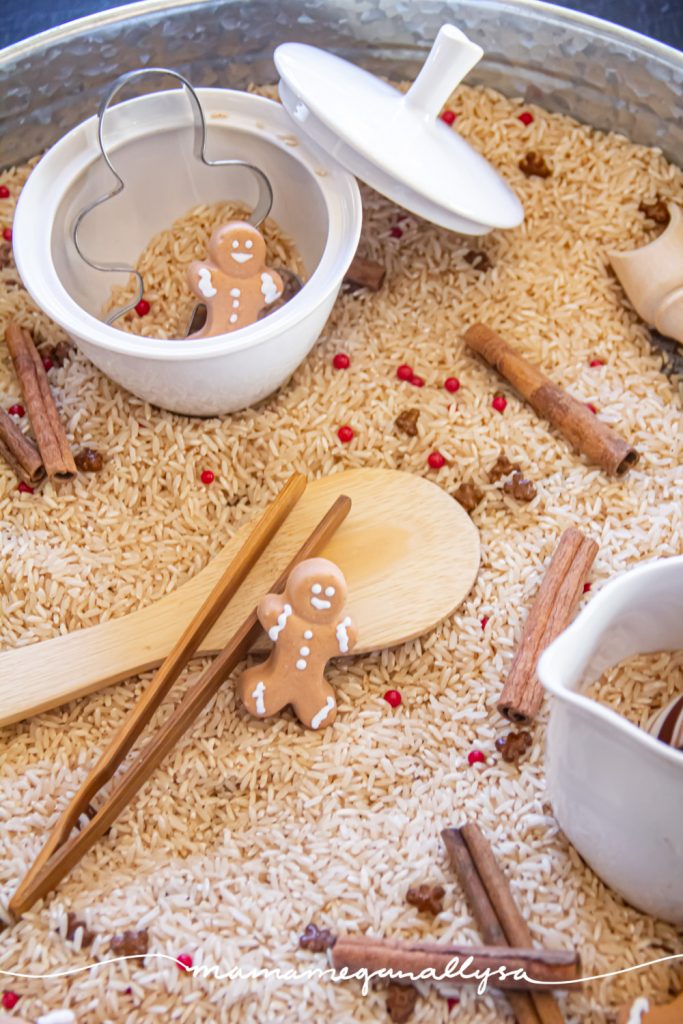 a detail shot of the set up in our gingerbread sensory bin. Using rice sented as gingerbread as the filler and sprinkles and cinnamon sticks mixed in. There are also gingerbread man candies scattered around as well as a gingerbread man cookie cutter and a couple other little tools and a creamer and sugar bowl for pouring