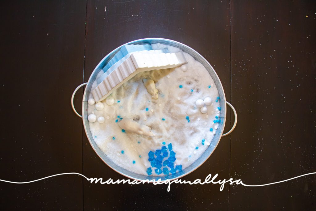 an arctic small world sensory bin filled with Epsom salt, a wooden ice cave stacker standing in the back of the tray with a mama polar bear and per cub stand in the shelter. Blue crystals and "snowballs" are scattered around