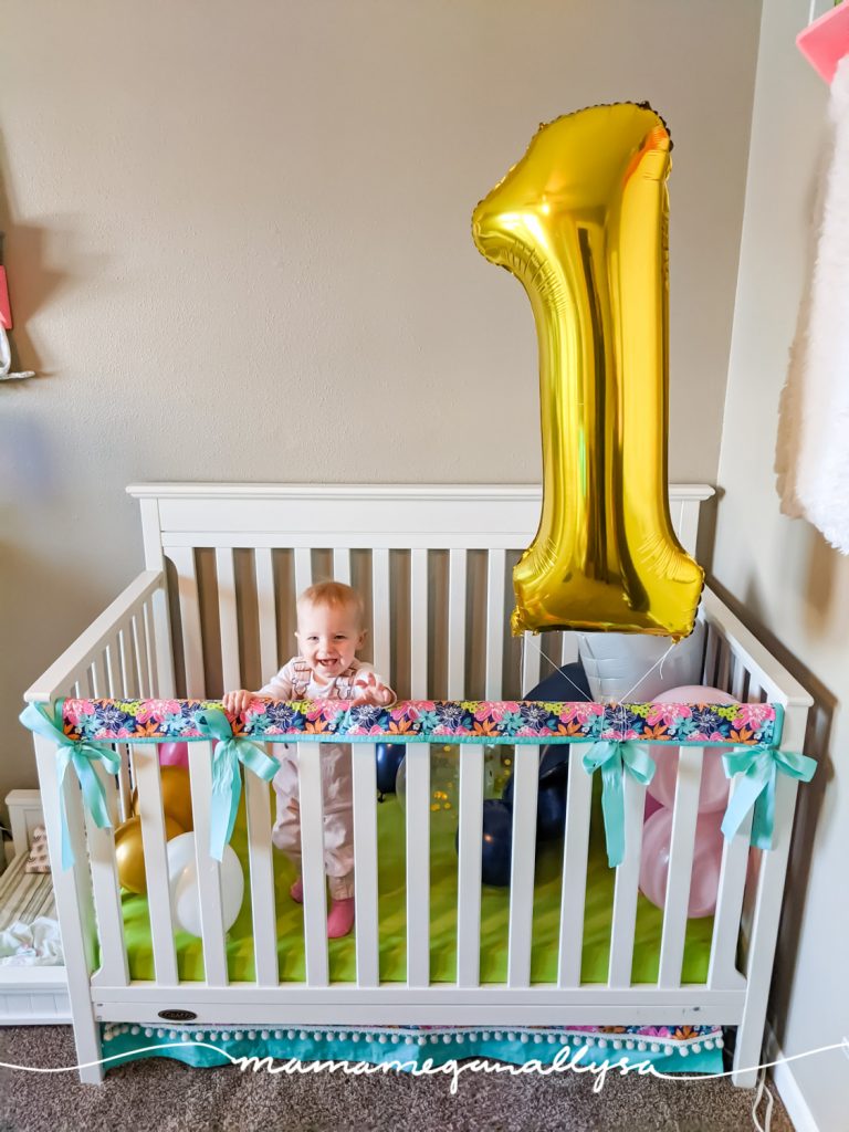 a baby girl standing in her crib that is filled with balloons and a big gold 1 balloon tired to the side