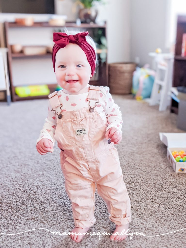 a baby stnading in pink overalls and a maroon headband