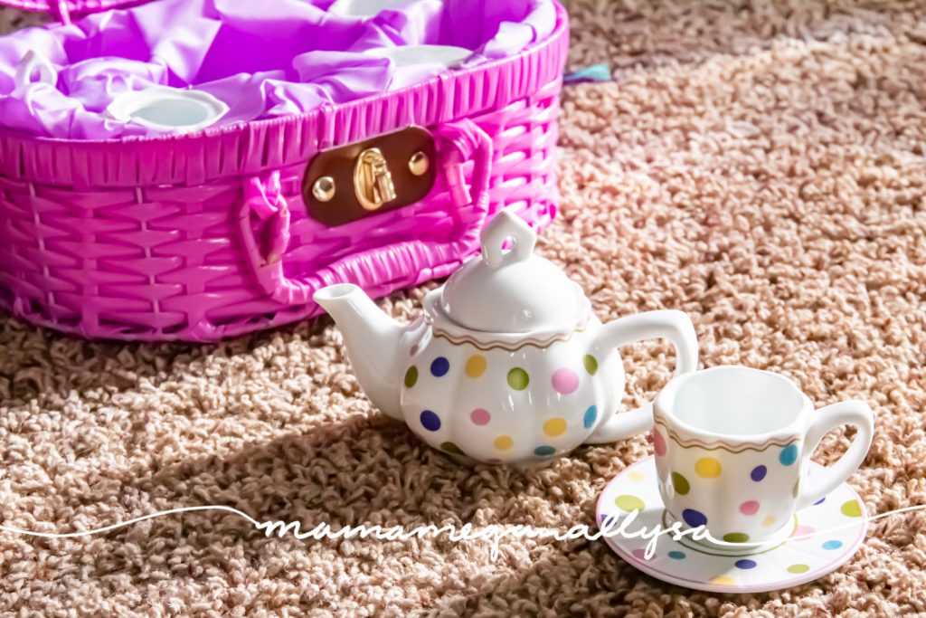 a close-up shot of a little kids polka dot porcelain teapot and cup on a saucer in front of a purple wicker carrying case