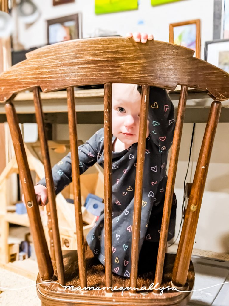 a little girl peeking through the back of a child sized wooden rocking chair