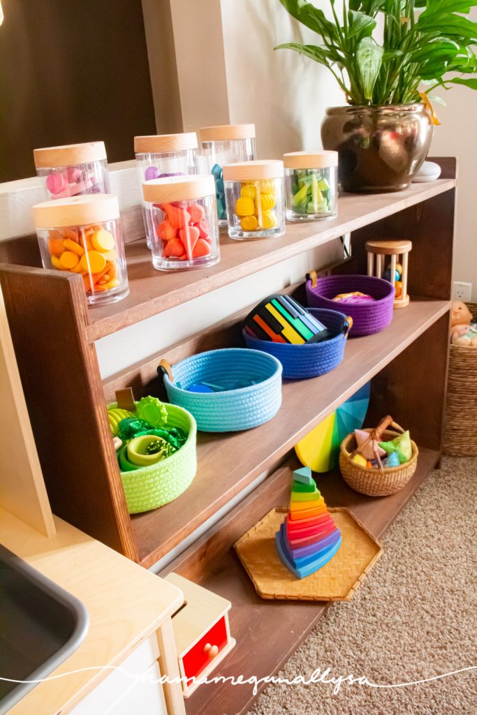 a toy shelf in our livingroom playroom, a rainbow of loose parts jars on the top shelf, colorful rope baskets on the second and other rainbow toys on the bottom shelf