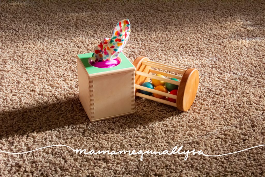 a couple of baby toys on the floor a Lovevery tissue box and a wooden cylinder rolling rattle