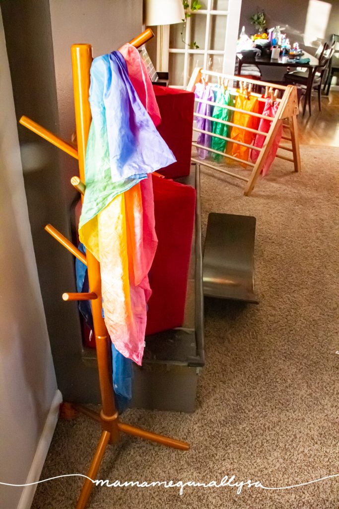 a kids tree stand with our rainbow play silk hanging on it with our pikler triangle in the background with the rest of our play silks  hanging