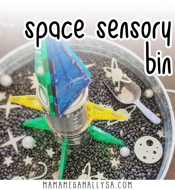 space sensorybin with black beans as the filler some glittery silver asteroids, glow in the dark stars and planets. In the center is a rocket made of tin cans and magnatiles