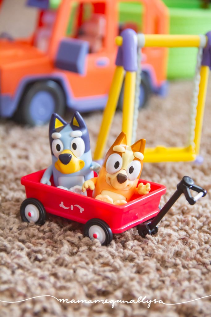 Bluey and bingo figures sitting in a red wagon in front of a swing and the bluey family car