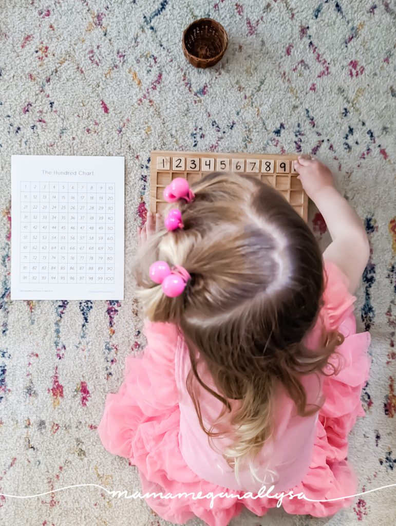 a little girl all dressed in pink putting wooden number tiles along the top row of a wooden 100s board with a printed control chart next to her on the floor